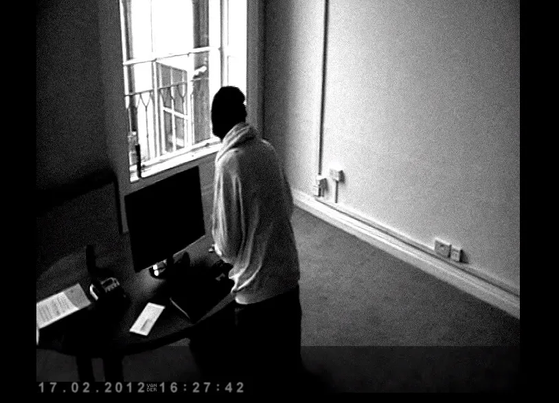 Black and white CCTV still, timestamped 2012, of a hooded figure standing at a desk in an office.